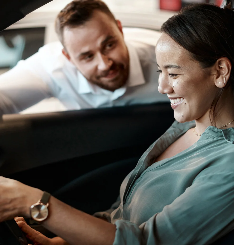 A happy customer sitting behind the wheel of her new car talking to the sales man