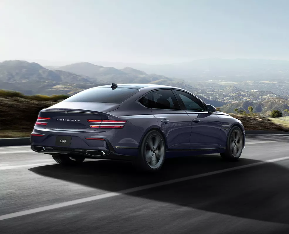 Back side view of a 2027 Genesis G80 driving on a road with mountains in the background