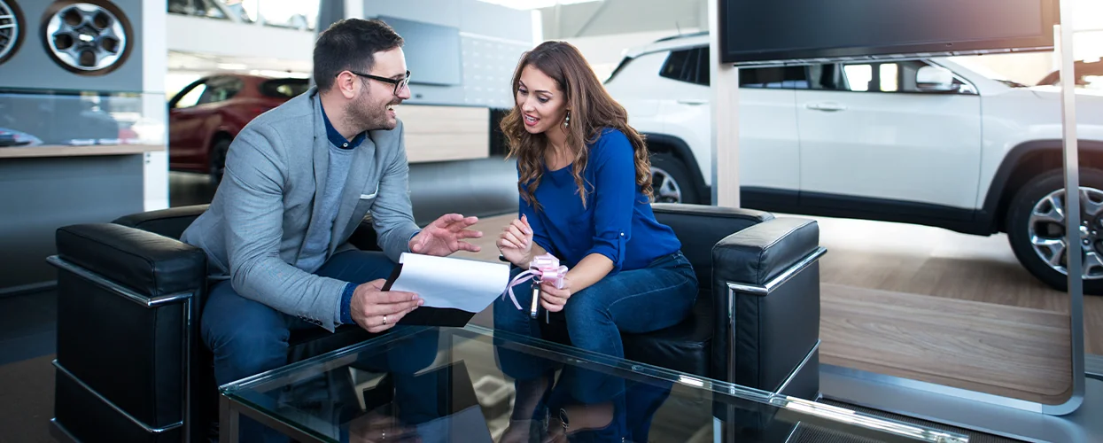 A man and woman in a car dealership, reviewing car models while seated in a comfortable waiting area.