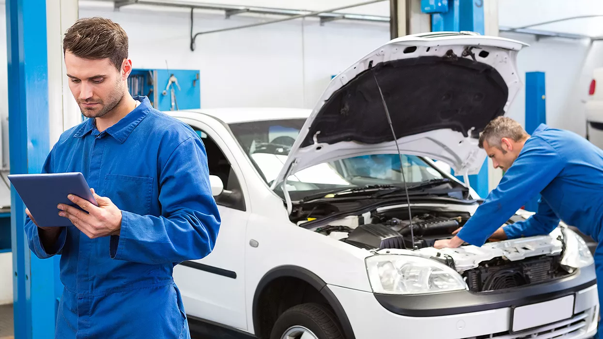 One mechanic looking at a cipboard while another works under the hood of a car