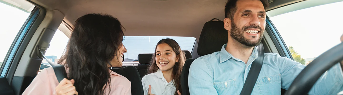 A family driving in their vehicle. Husband at the steering wheel smiling, wife in passenger seat looking back at their daughter sitting in the middle seat smiling.