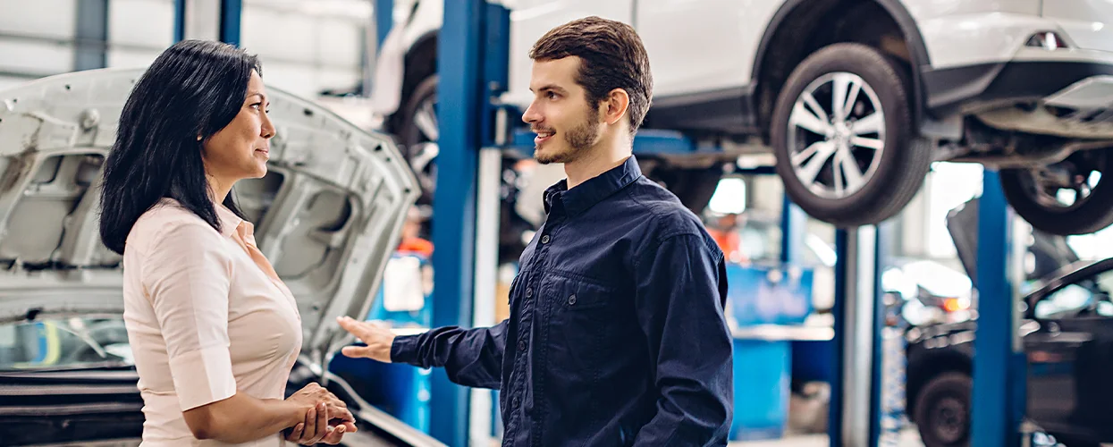Inside a car repair shop, a man and woman engage in conversation, with various car parts visible around them.