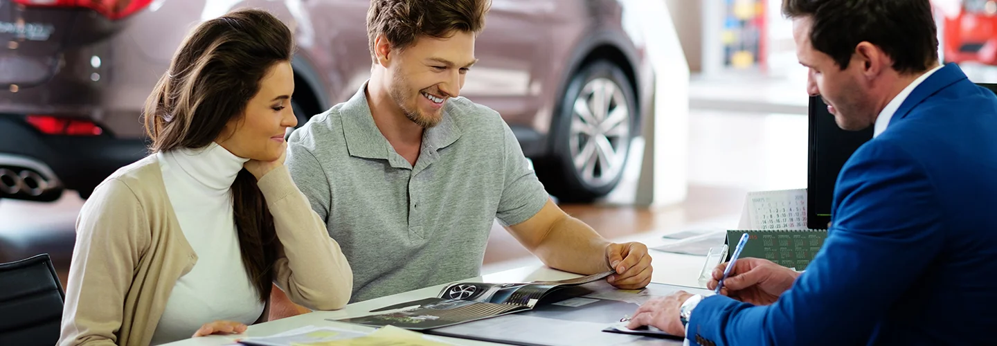 Inside a car dealership, a man and woman sit at a desk, engaged in conversation about purchasing a vehicle.