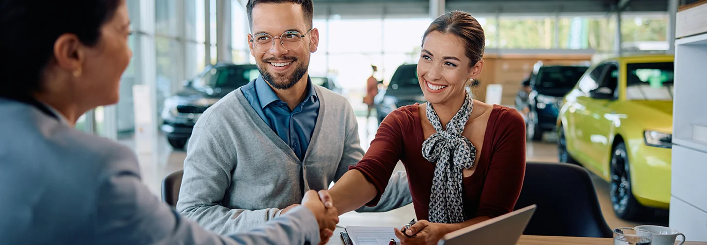 A man and woman shaking hands outdoors, with a car behind them, suggesting a meeting or negotiation context.