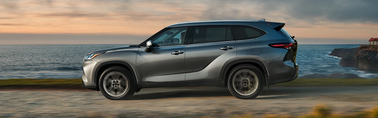 driver side view of a 2025 Toyota Highlander Hybrid XLE driving on dirt road by water