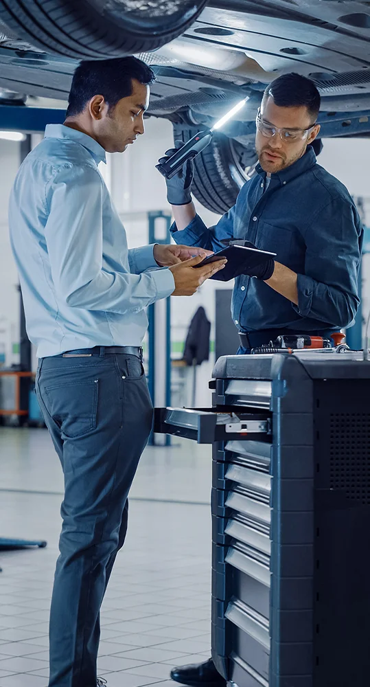 Two men in a garage focused on a tablet, with automotive tools and parts in the background, indicating a work-related task.