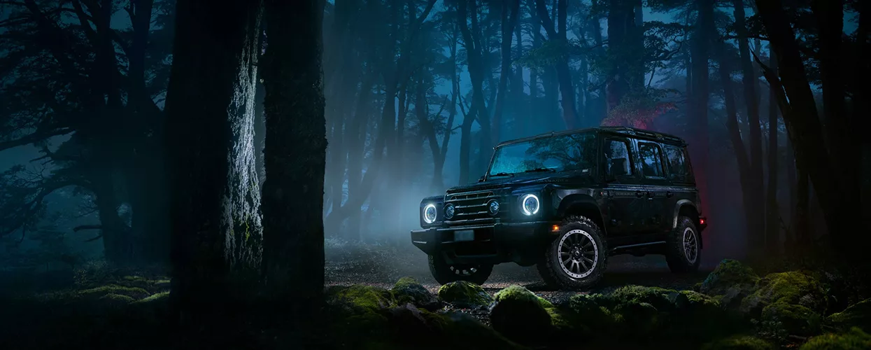 a black INEOS Grenadier Black Edition driving with lights on in forest at night