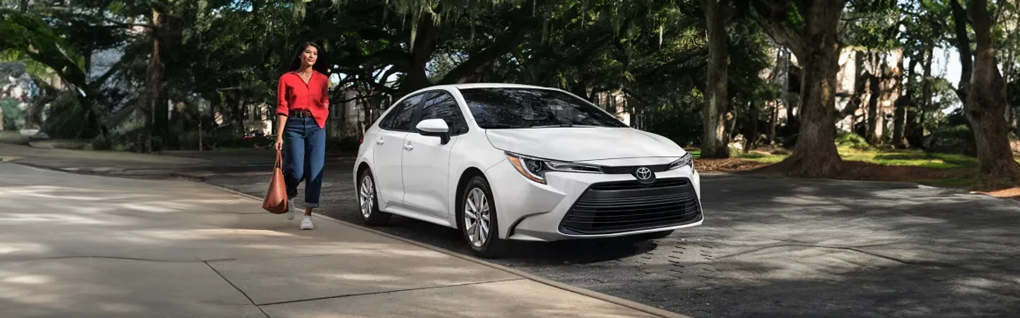 A white 2024 Toyota Corolla parked on the street with a women talking to it