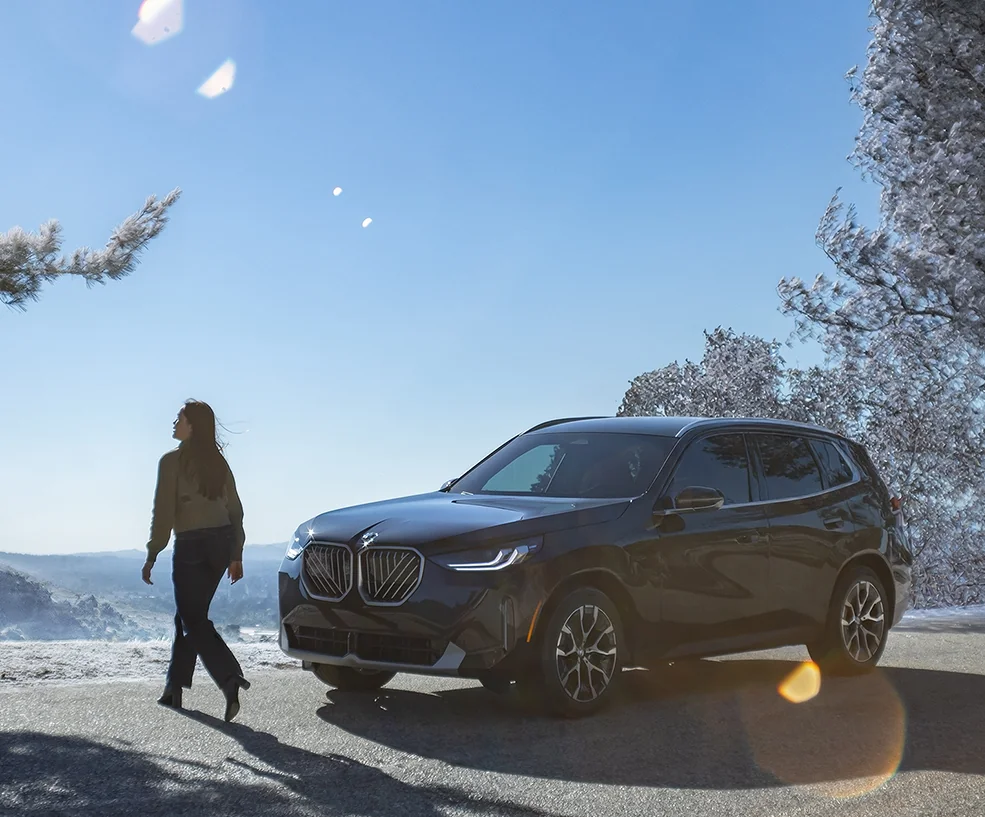 Front side images of a black 2026 BMW X5 parked next to some trees with snow on them