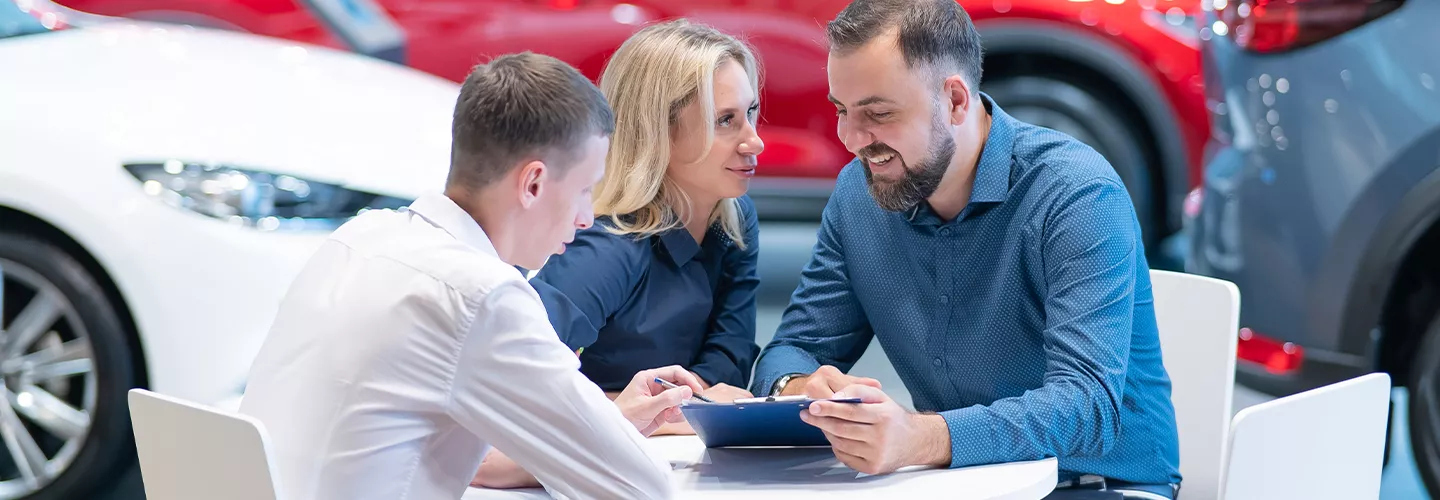 Car salesman sitting with two people at at table in a car dealership