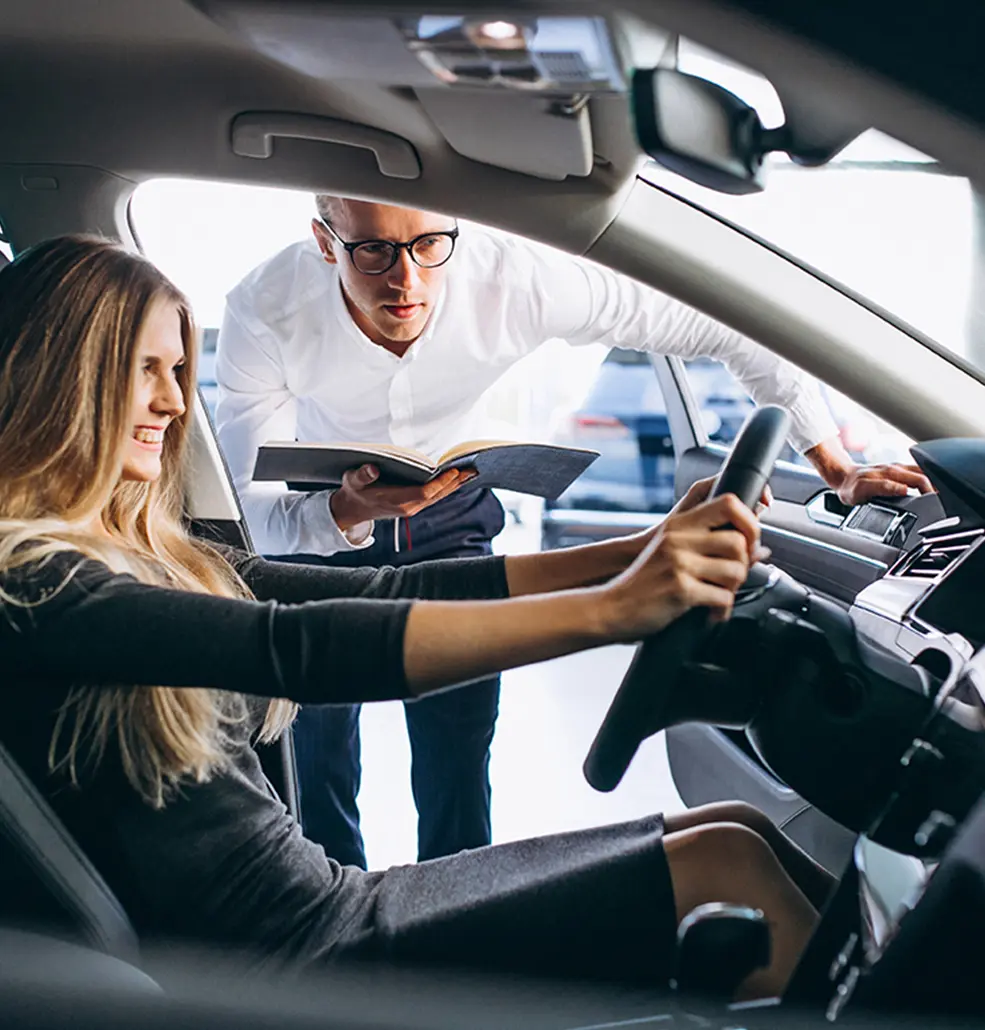woman smiling sitting in new car on lot while rep holds owners manual