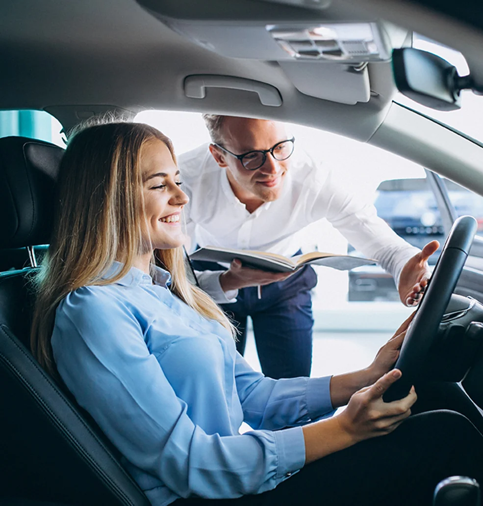 Inside a car, a man and woman gaze at a nearby vehicle, discussing its features.