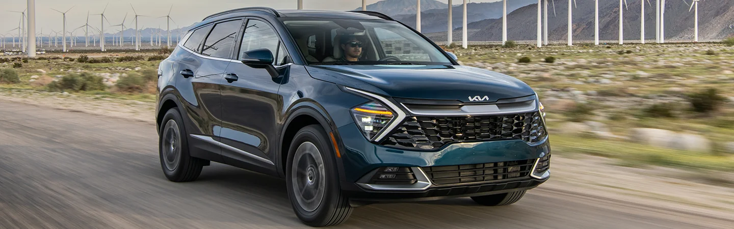 Front view of a 2025 Kia Sportage Hybrid driving on a road next to a wind farm