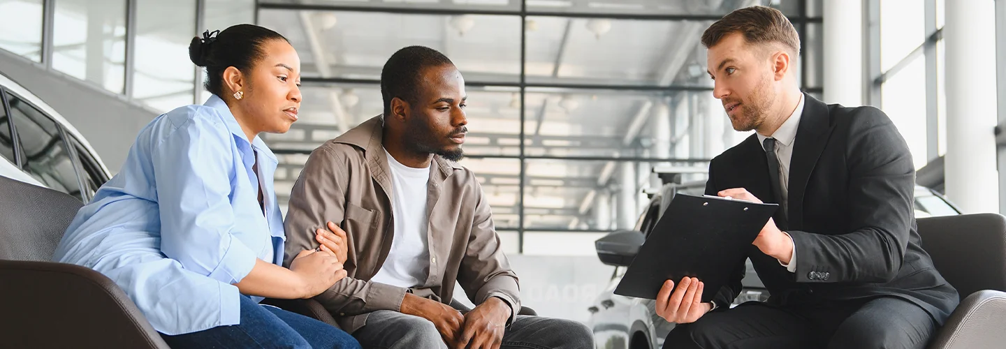 Three people engaged in conversation while seated in a car dealership, surrounded by vehicles and promotional materials.
