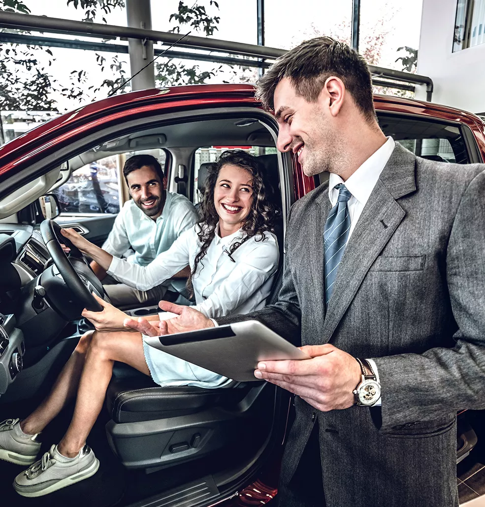 Couple sitting in a new vehicle with a car salesman standing outside
