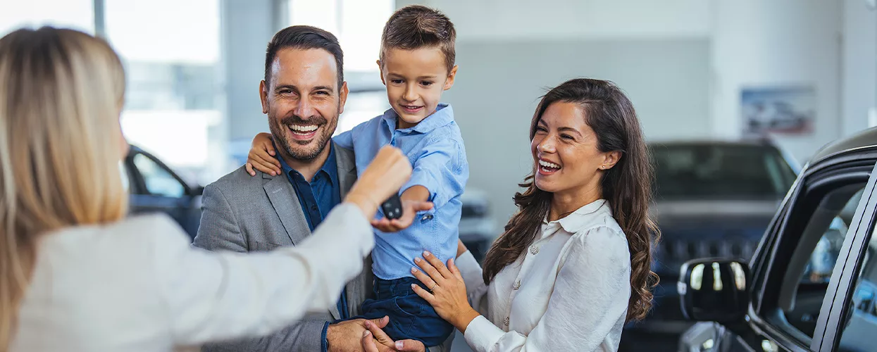 sales woman handing the car keys to the child that the father is holding and mother standing next to them.