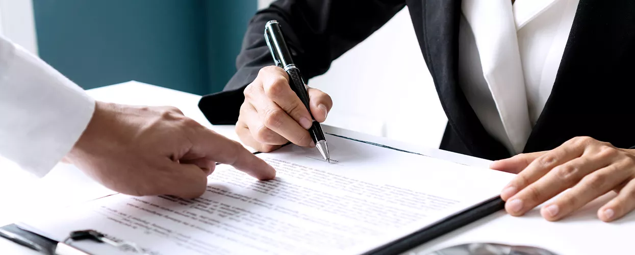 Person signing a document with another person pointing where to sign