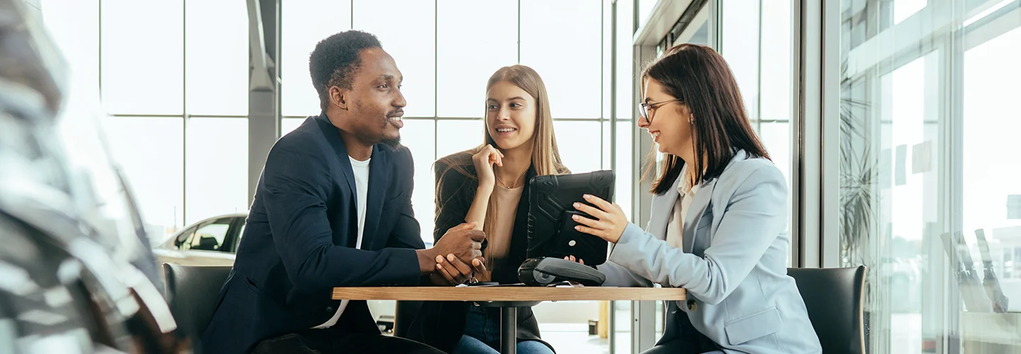 couple in dealership showroom discussing a car purchase with salesperson holding a tablet pc showing information
