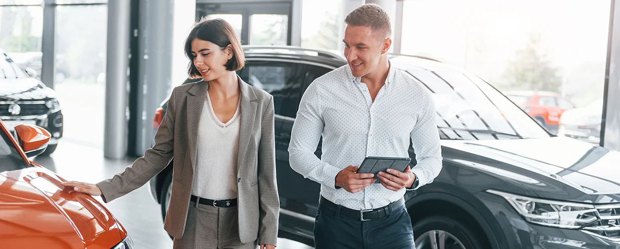 A man and woman pose in front of a car, both smiling and wearing casual clothing.