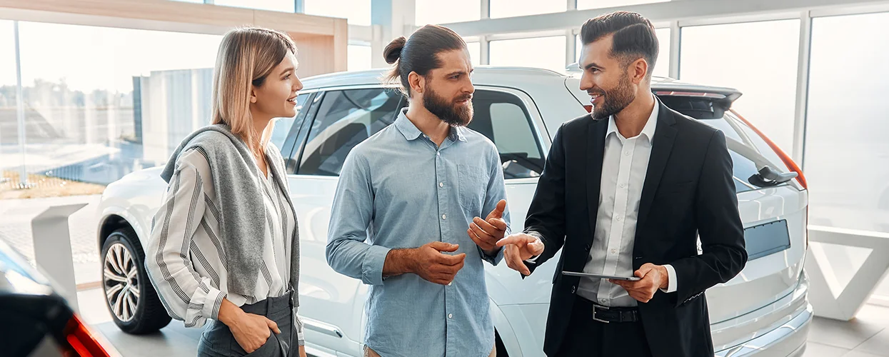 Two people consult with a salesperson in a car showroom, exploring vehicle choices and features.
