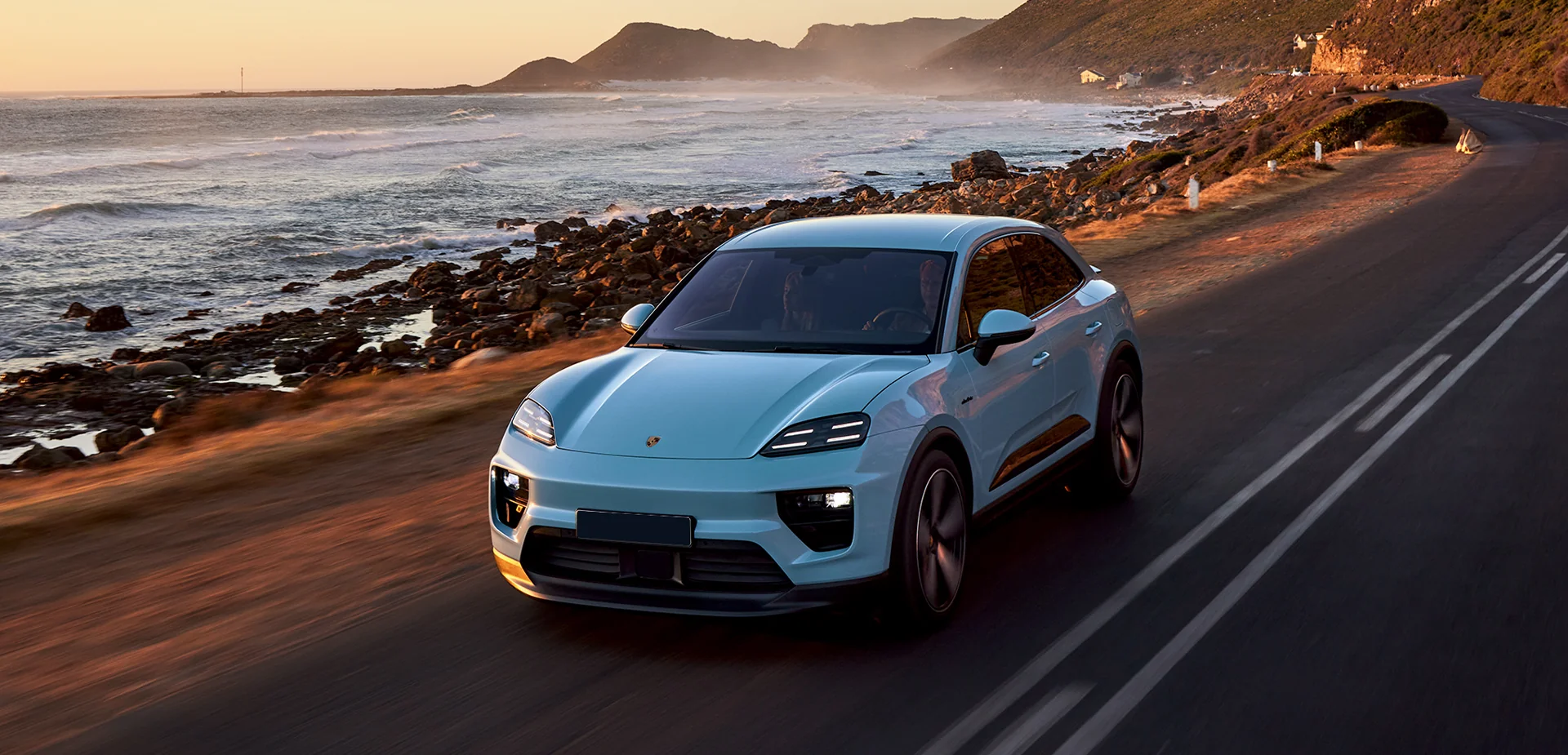 Front view of a powder blue 2026 Porsche Macan driving on a road by the ocean