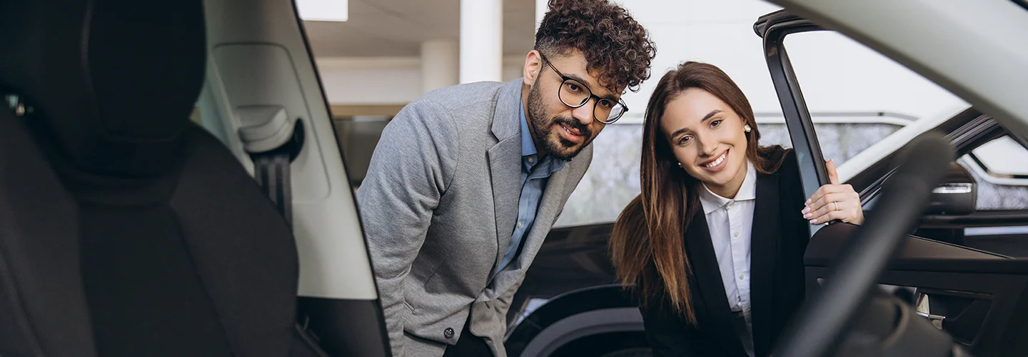 A man and woman stand side by side, looking intently at a car, possibly considering a purchase.