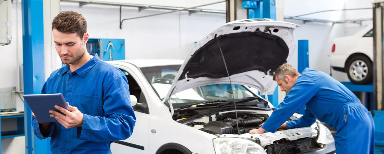 Two service mechanics, one working under the hood of a car and the other holding a clipboard