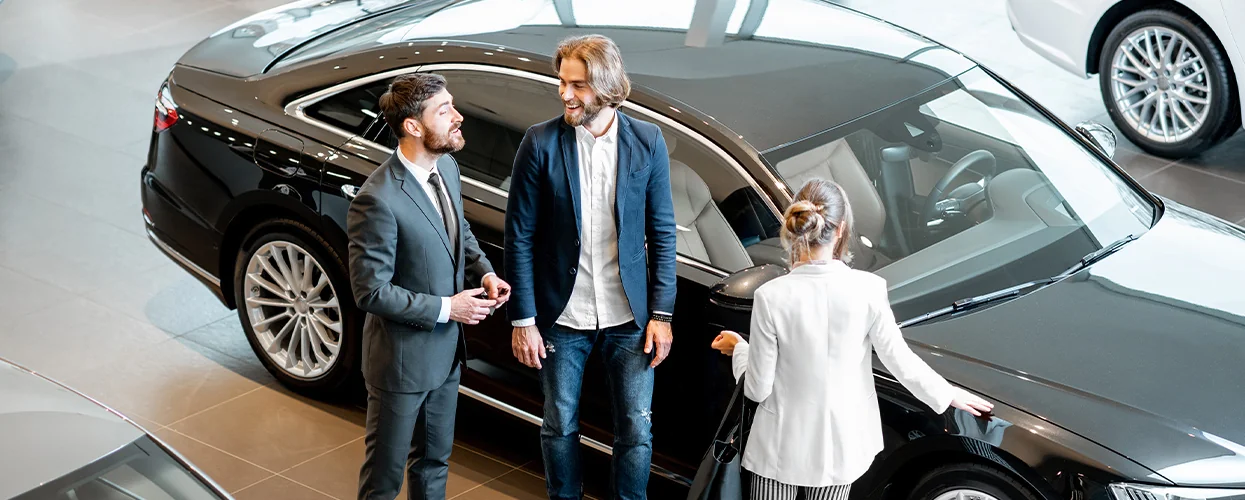 Three individuals stand around a car in a brightly lit showroom, discussing its features and specifications.