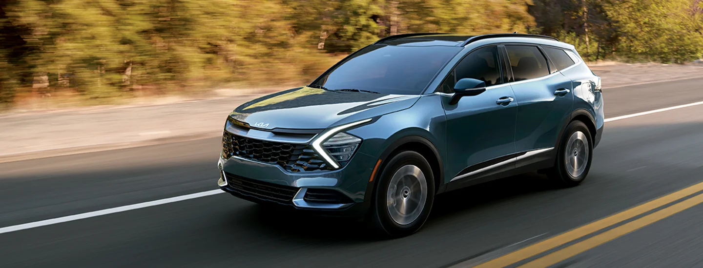 Front side view of a teal 2025 Kia Sportage Hybrid driving on a road next to some woods