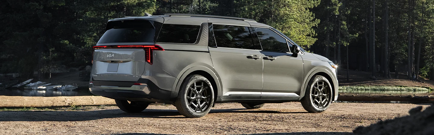 Back side view of a parked gray 2025 Kia Carnival Hybrid by a lake