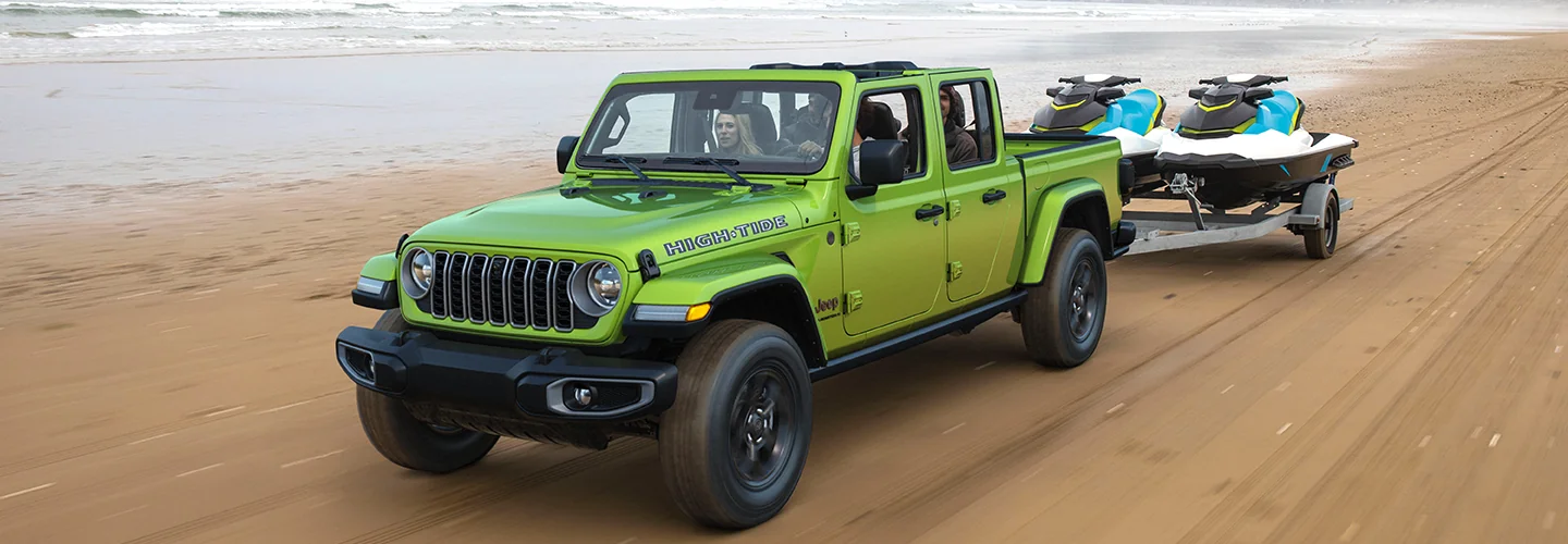 A green jeep is pulling two jetskis on the beach, with waves gently lapping at the shore.