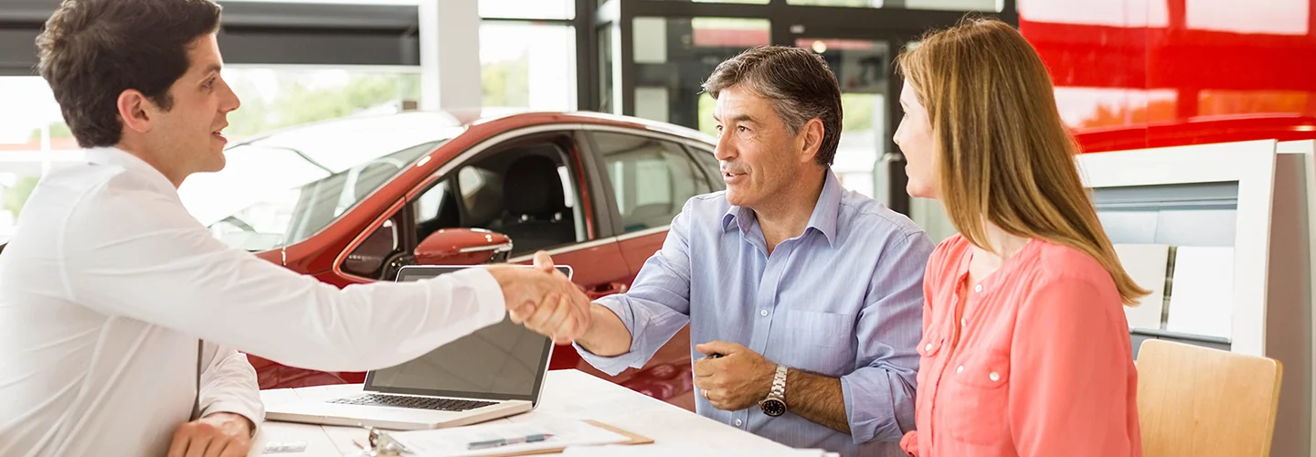 A man and woman shake hands in front of a parked car, symbolizing a business agreement or partnership.