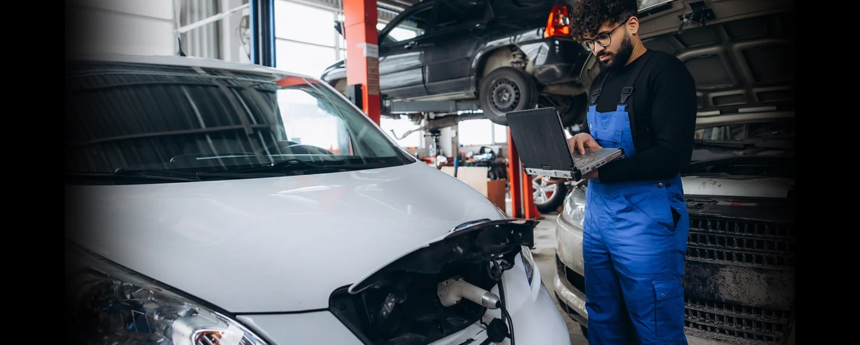A man wearing blue overalls is next to a car, working on a laptop, likely for automotive troubleshooting.