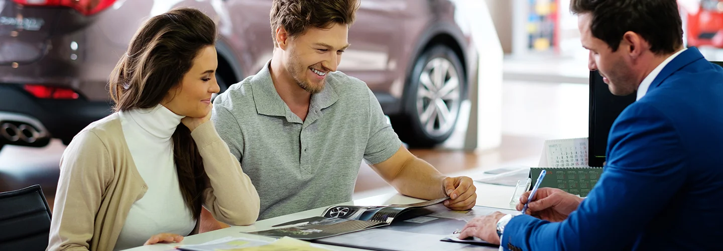 A man and woman are seated at a table in a car dealership, reviewing vehicle options and paperwork together.