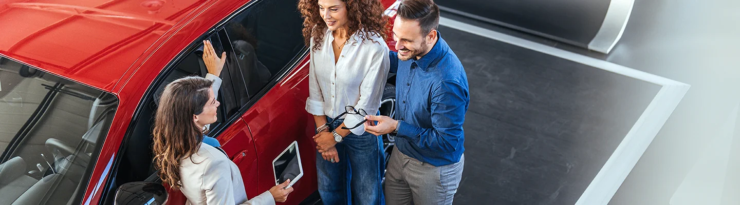 saleswoman talkign with husband and wife next to a red vehicle
