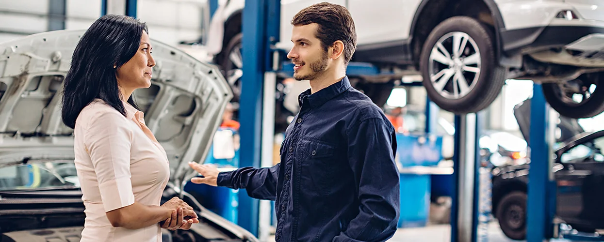 A man and woman discussing car repairs in a busy auto shop, with various tools and vehicles in the background.