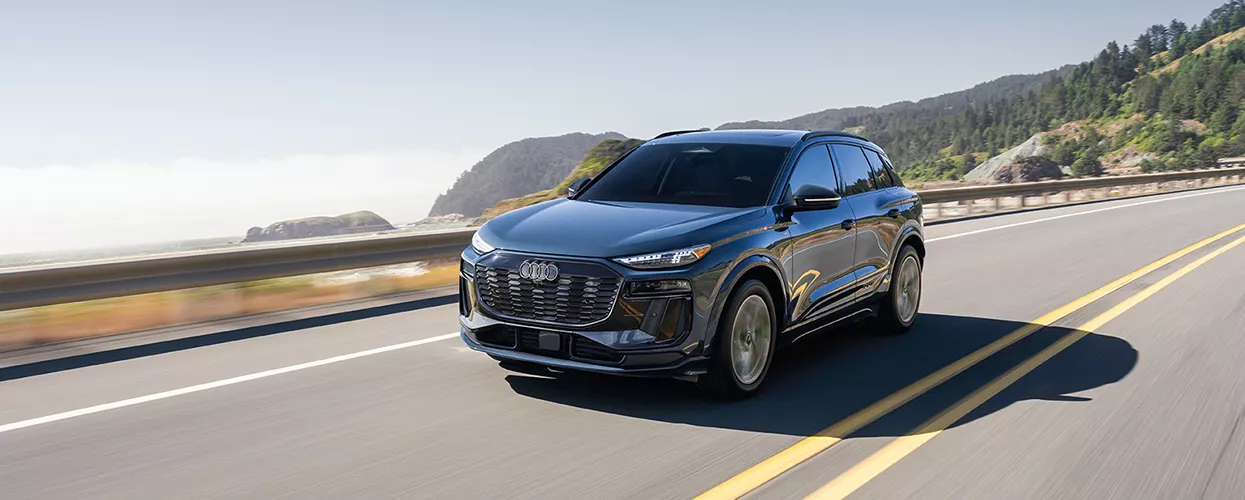 an Audi Vehicle displayed driving on a coastal road with view of seashore as backdrop.