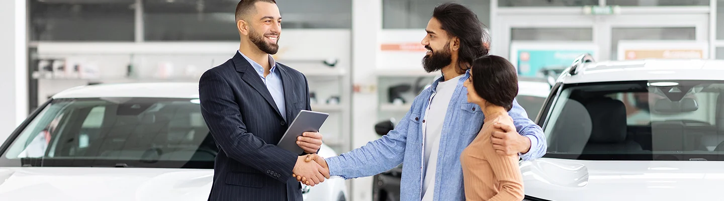 two people shaking hands with one along side in a car dealership making a new car purchase
