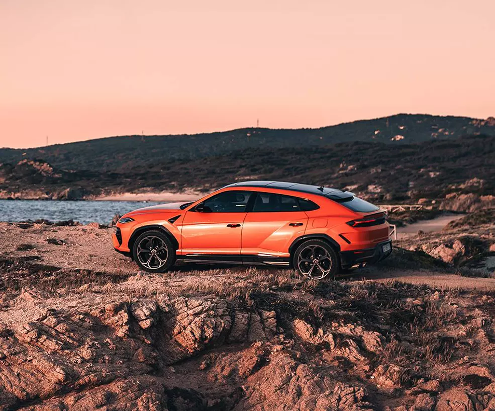 Side view of a orange 2026 Lamborghini Urus SE parked on a rocky ocean shore