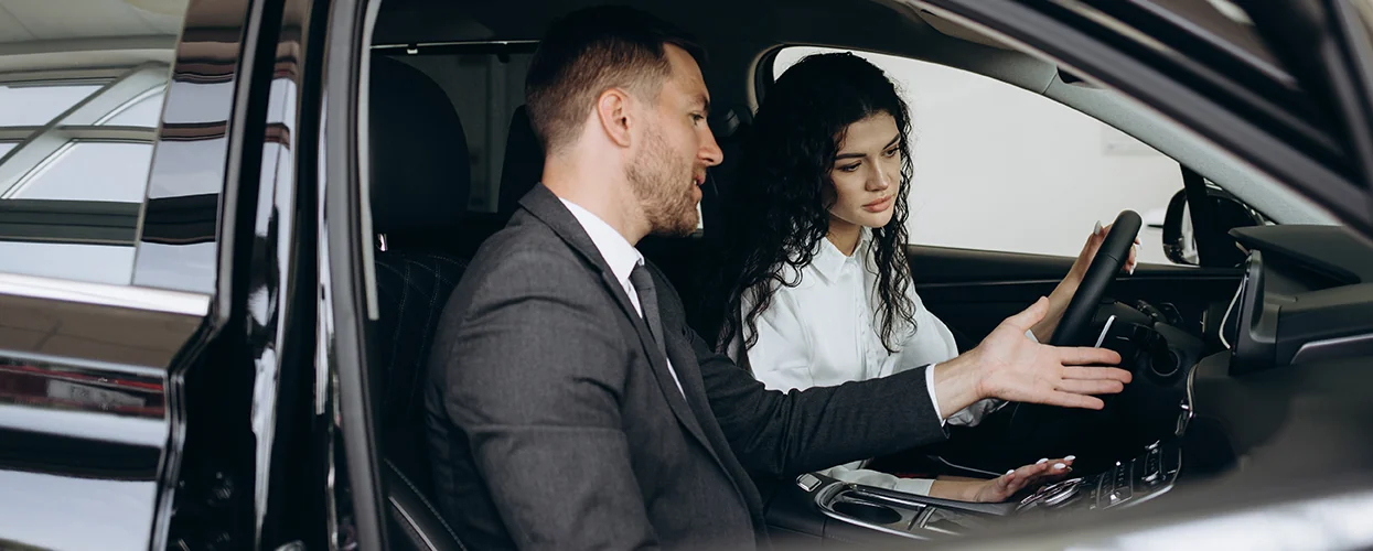 two people sitting in a car in the showroom one explaining the features of the car while sitting in it
