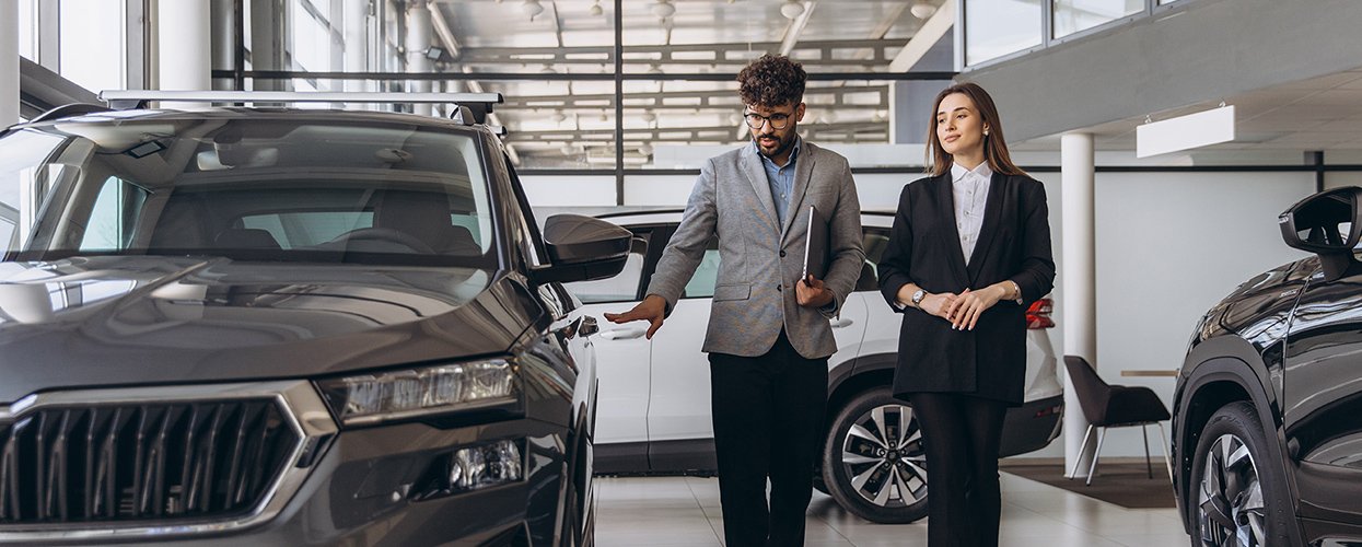 Two individuals stand beside a car in a showroom, discussing its features and specifications.