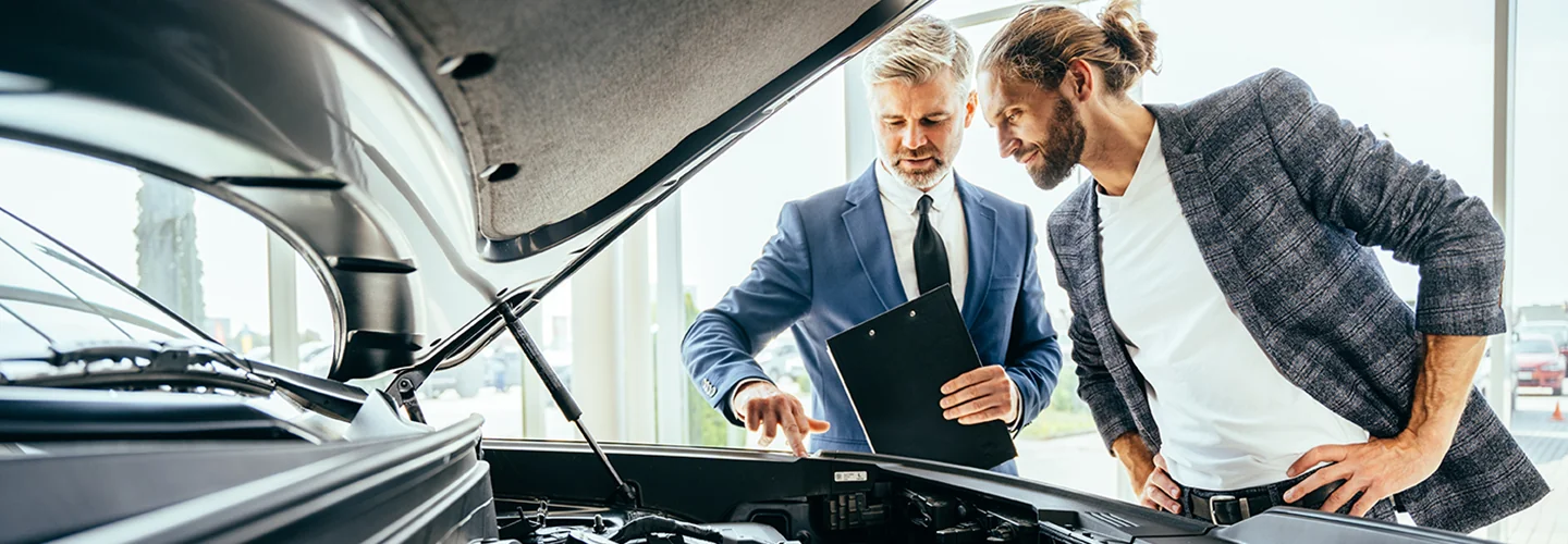 Two men examining a car engine, engaged in conversation about its functionality and maintenance.