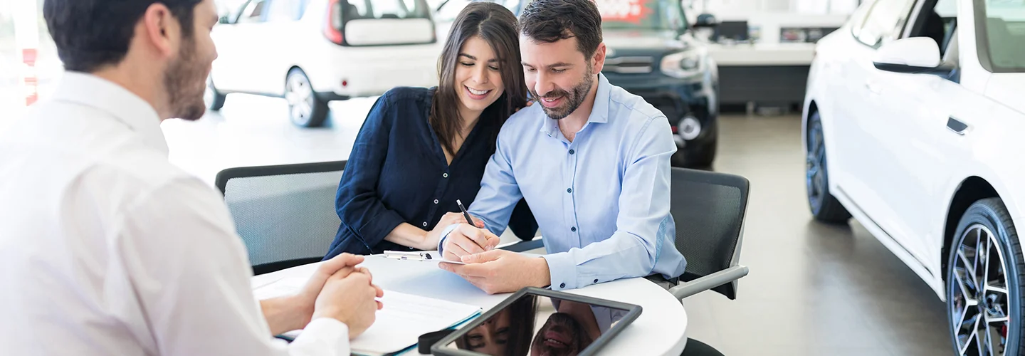 A man and woman sit at a table in a car dealership, discussing options with a salesperson nearby.