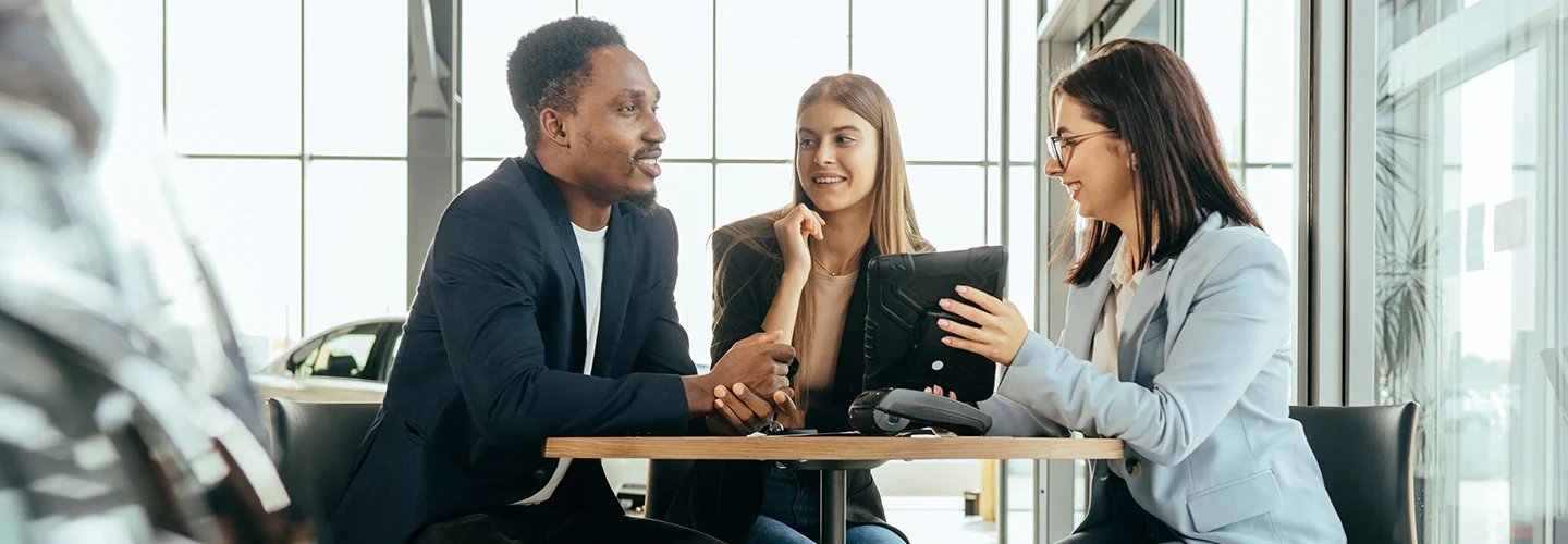 Two people seated at a table inside a car dealership, engaged in conversation about vehicle options.