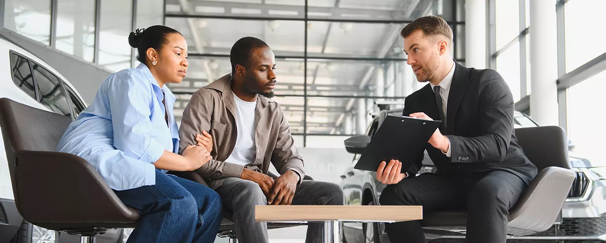 a couple sitting down with car salesperson in showroom going over details on a clipboard