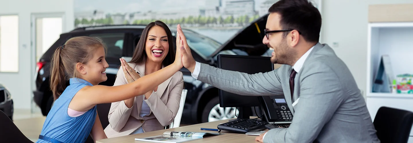 a salesperson is high fiving a child with another adult seated smiling about a car purchase