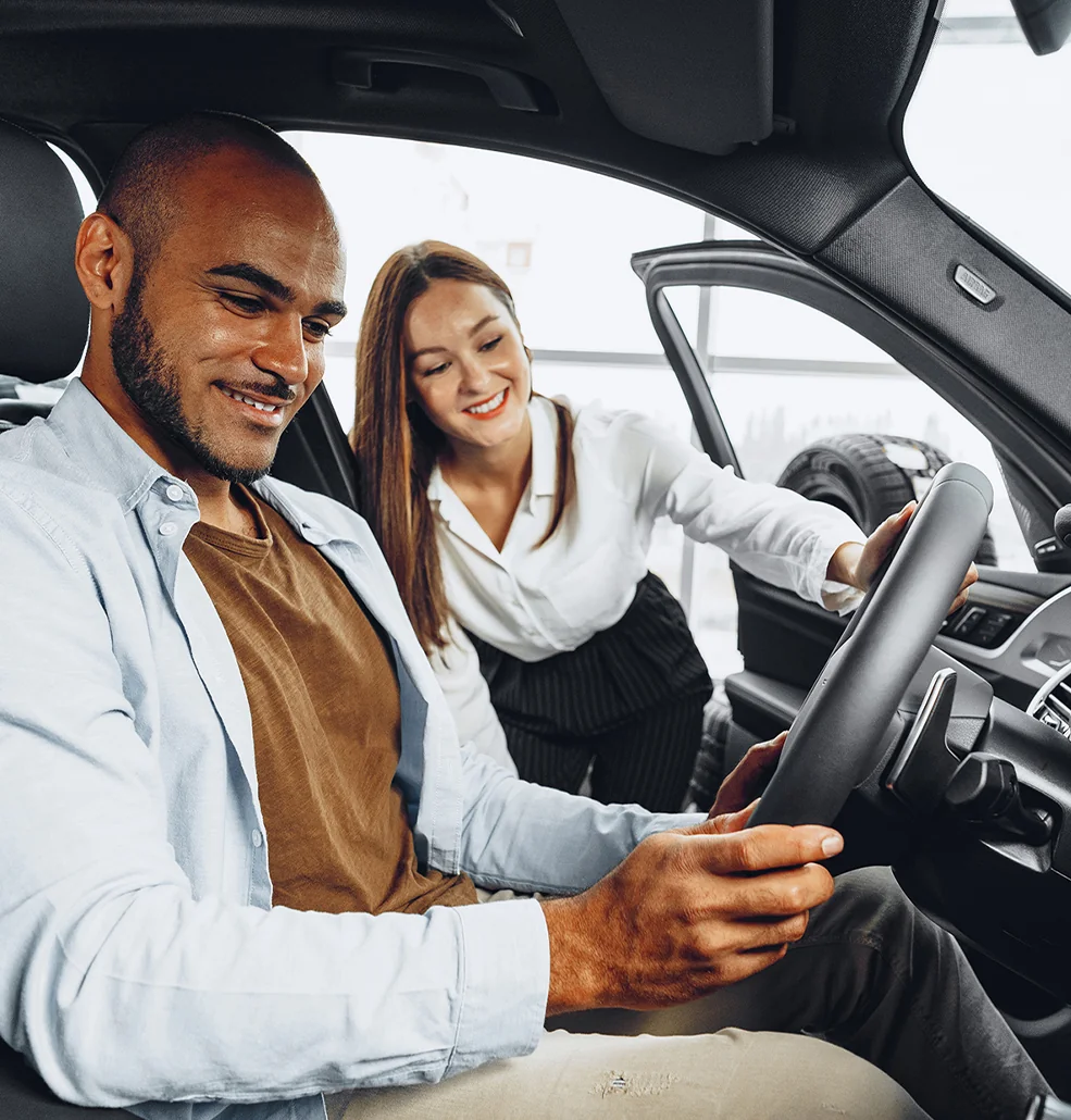 Inside a car, a man and woman gaze at a nearby vehicle, discussing its features.