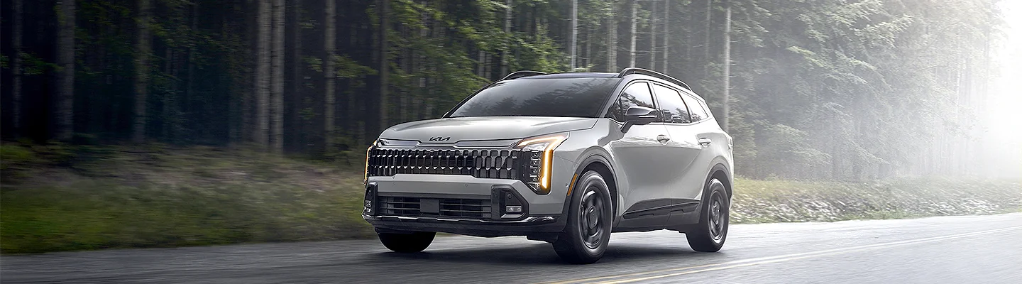 front view of a white 2026 Kia Sportage driving by woods