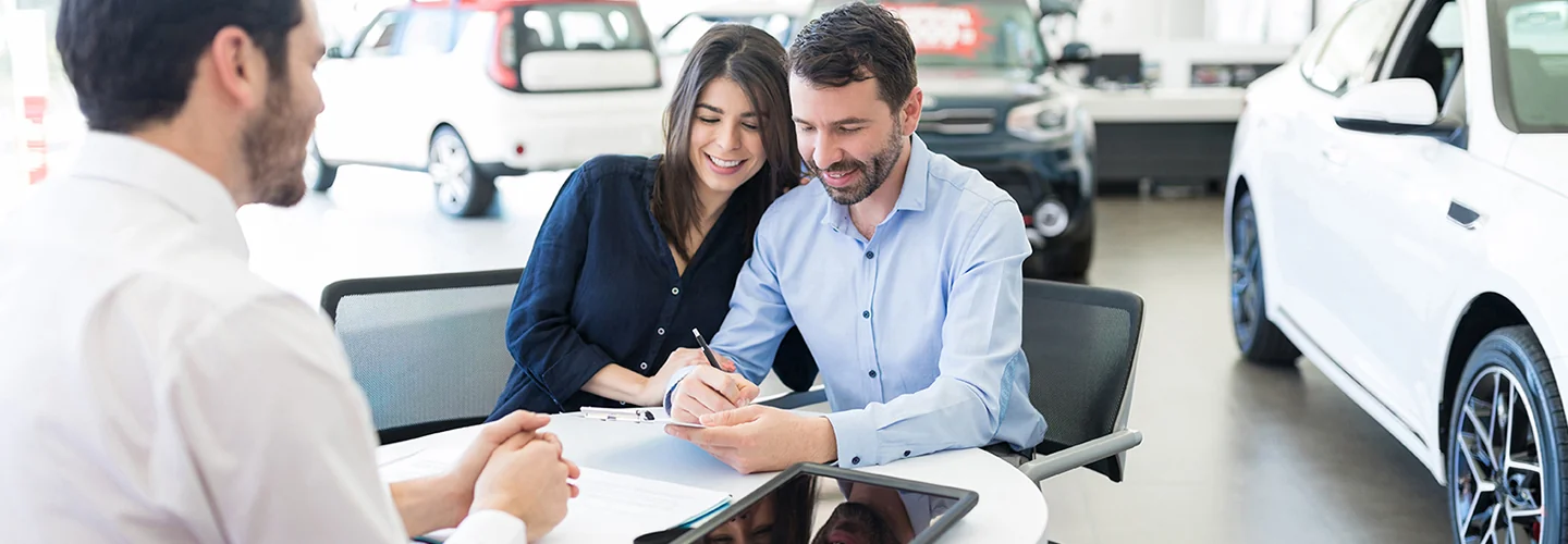 A man and woman seated at a table inside a car dealership, discussing options with a salesperson nearby.