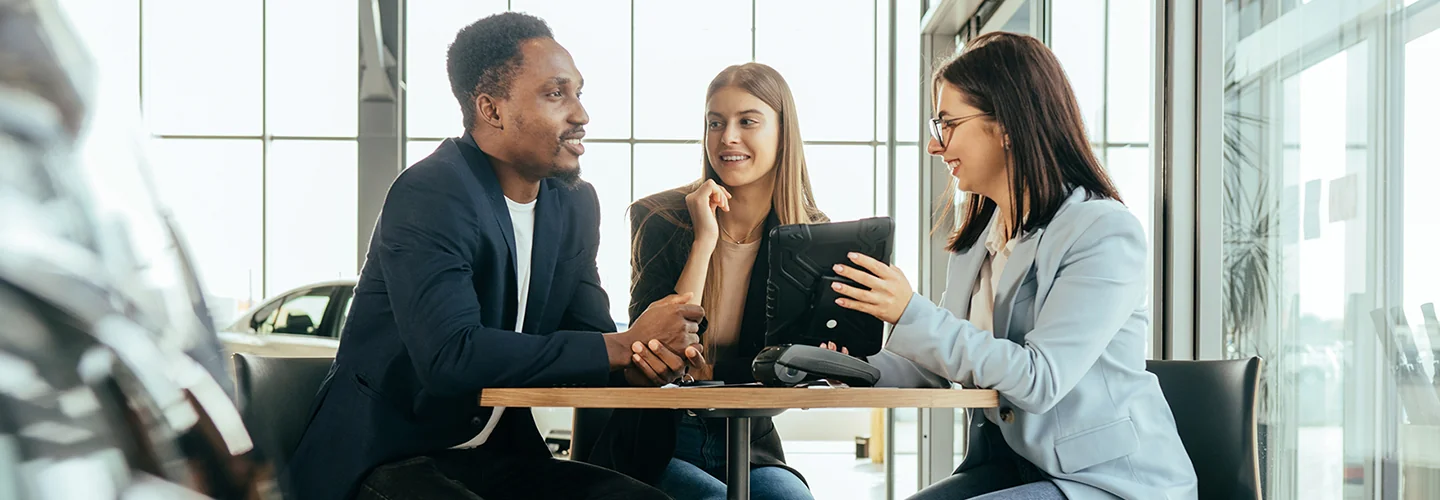 A man and woman sitting at a table in a car dealership, engaged in conversation about purchasing a vehicle.
